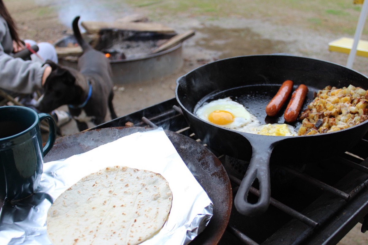 Utensilios de cocina de camping y verduras frescas sobre una mesa de madera al aire libre.