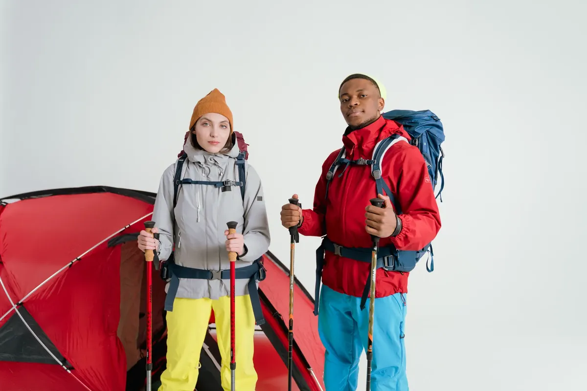 Two adults equipped with camping gear and hiking sticks pose in front of a tent on a white background.