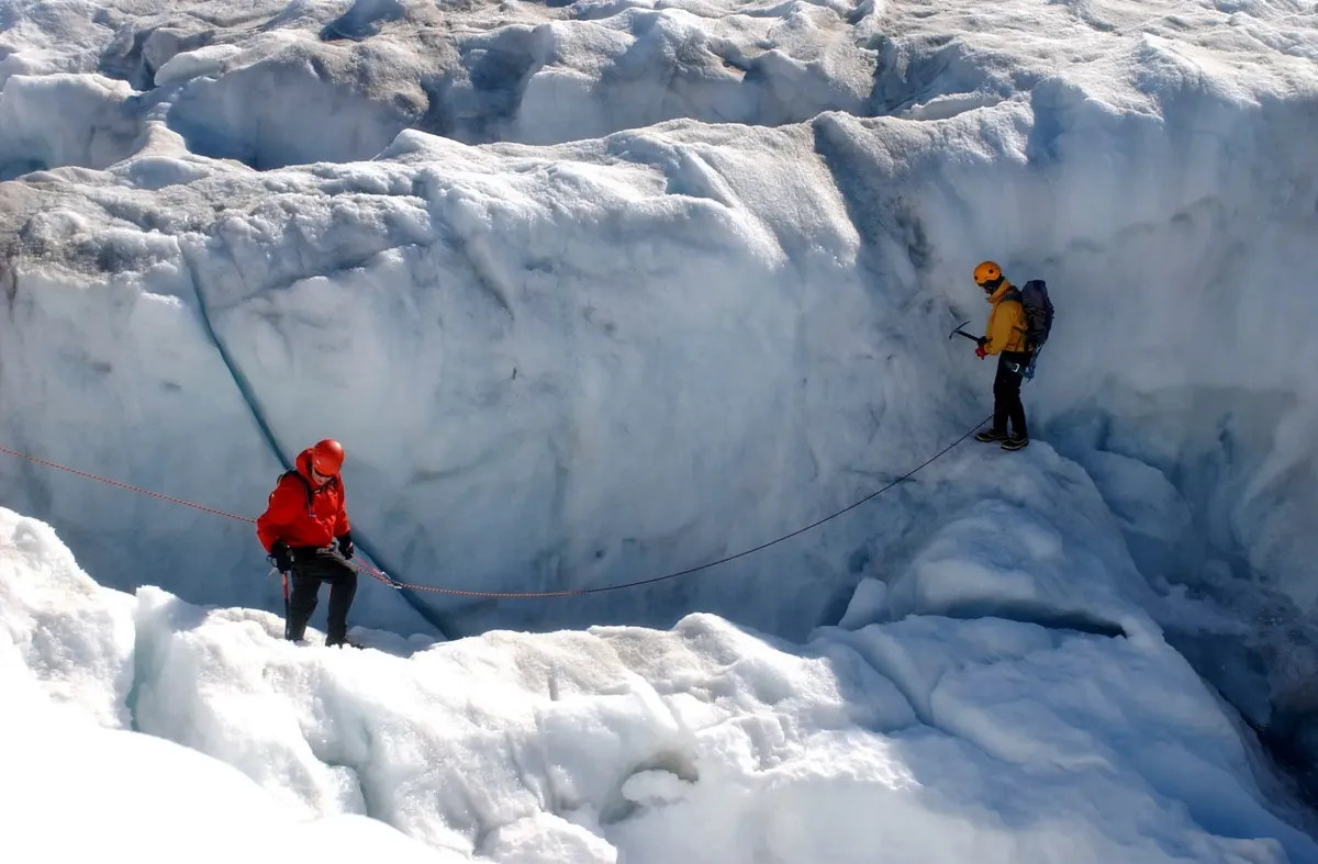 Two climbers navigate a glacier crevasse using ropes and ice axes in winter conditions.