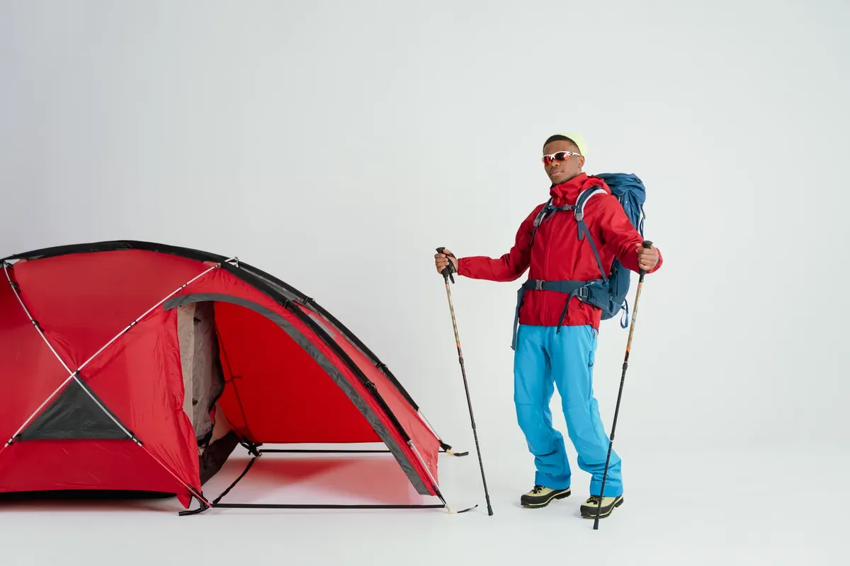 A man dressed for winter camping stands by a red tent with trekking poles.