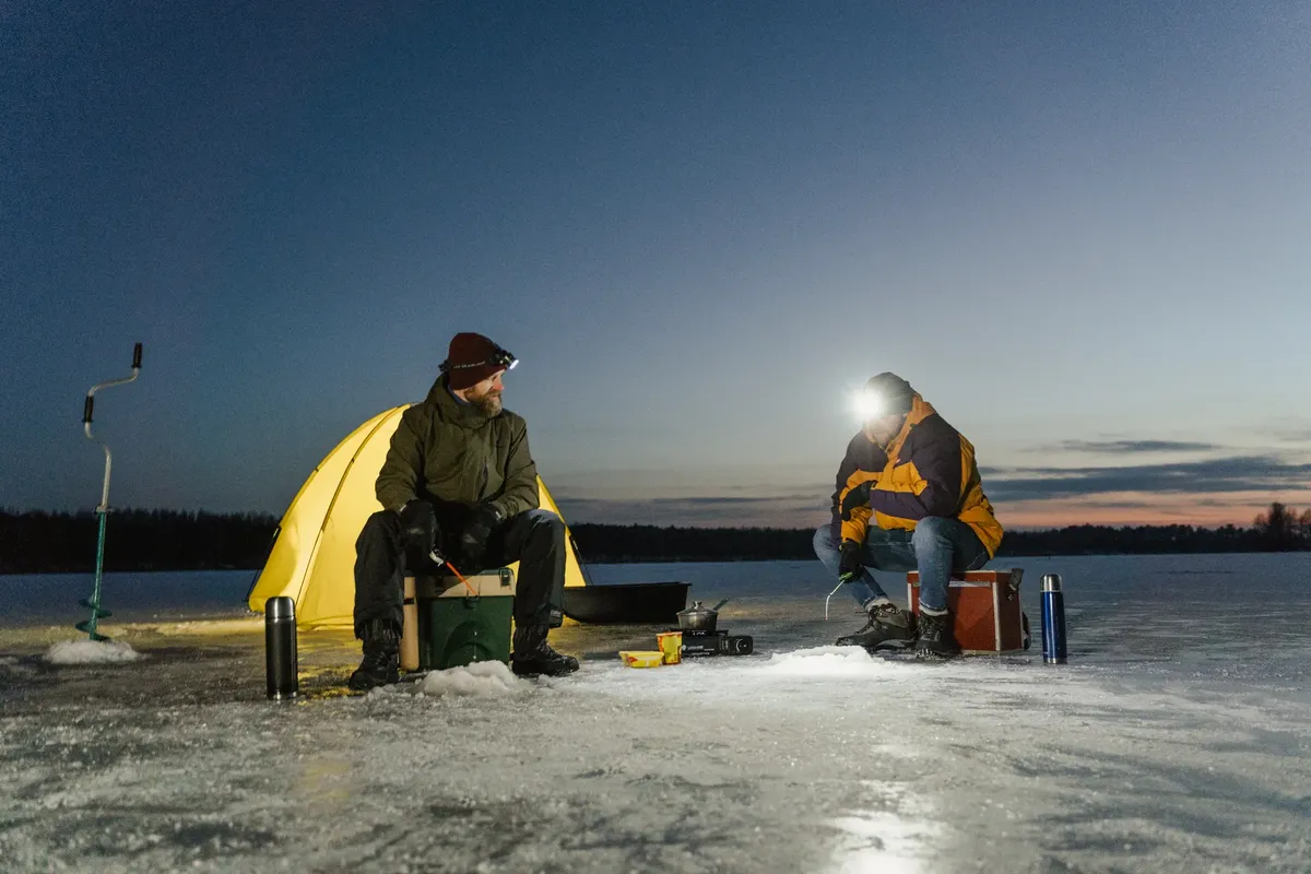 Two men ice fishing by a tent during winter dusk. Adventure on a frozen lake.