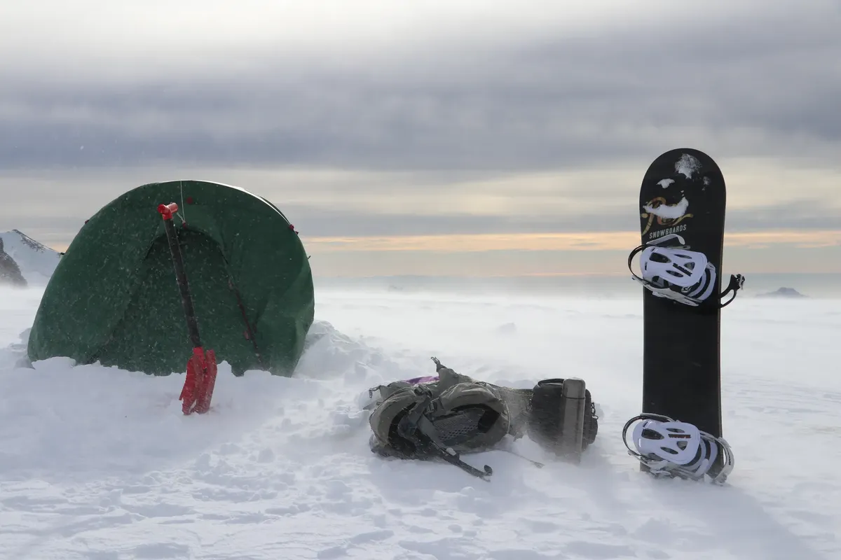 Snowy mountain camping setup with a tent, snowboard, and backpack in winter.