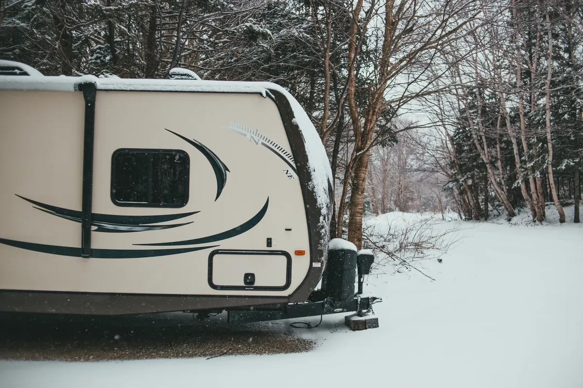 Camper trailer parked in coniferous and deciduous forest on road trip in snowfall in gloomy weather in winter in daytime