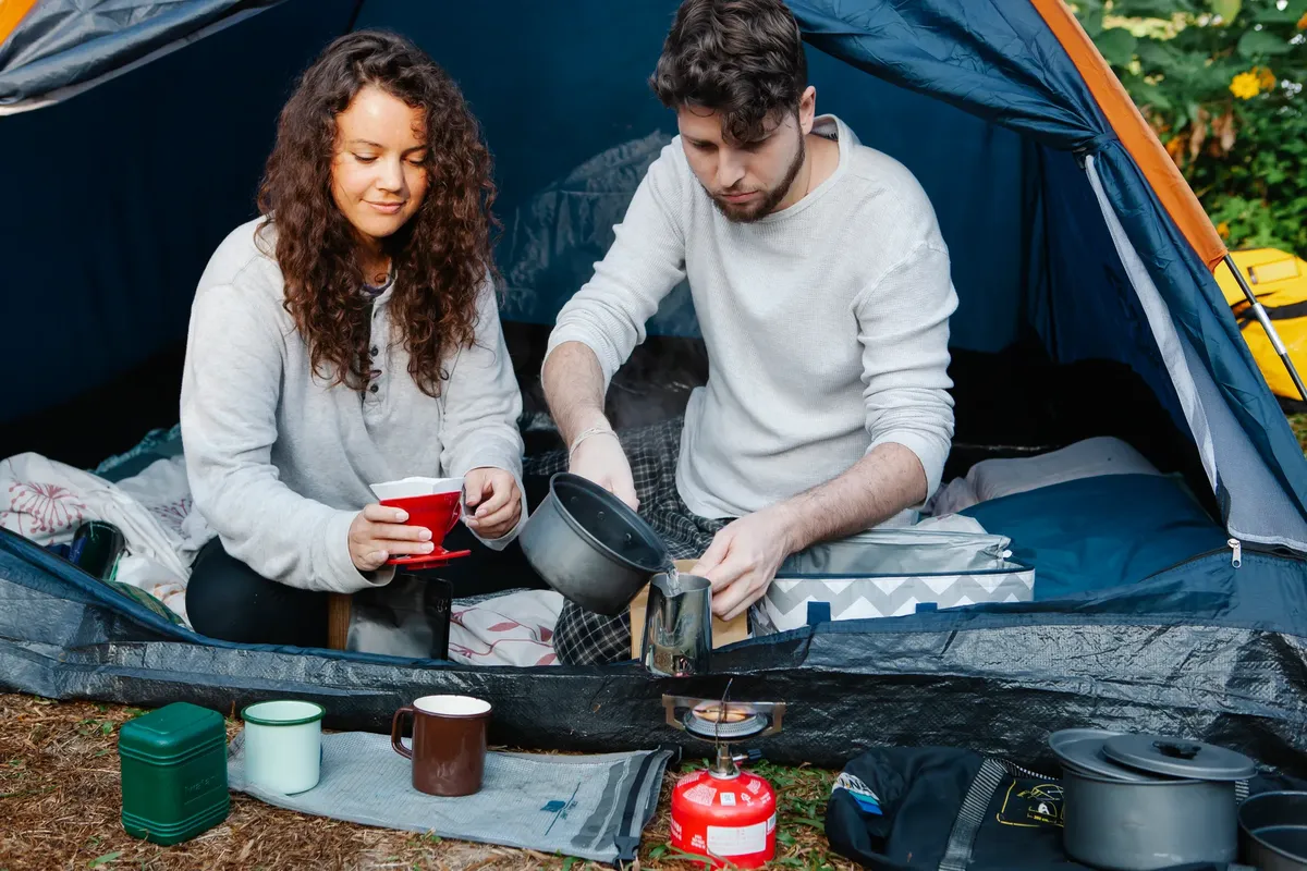 Crop young male tourist pouring hot water in pitcher near girlfriend with filter for coffee in campsite