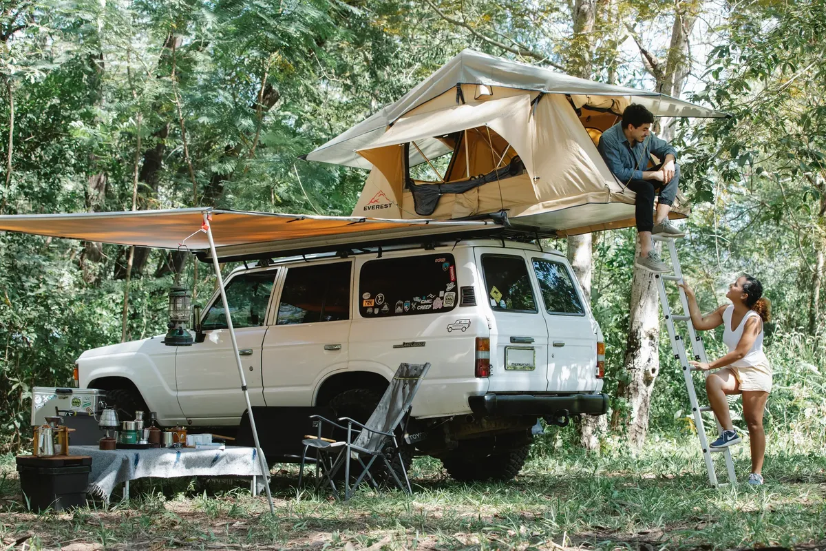 Full body of young male camper relaxing in tent placed on SUV car roof and chatting with Hispanic girlfriend climbing ladder during trip in nature on sunny day